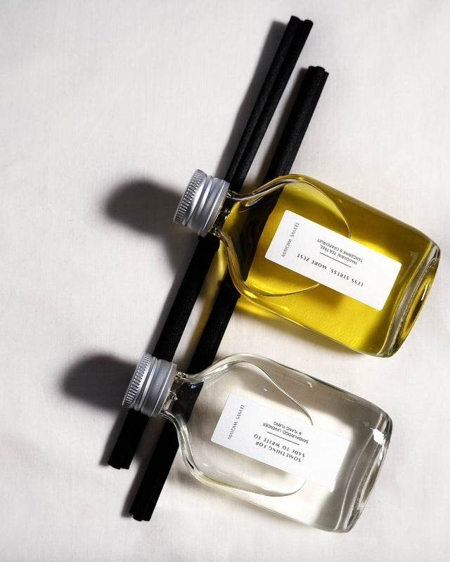 A minimalist overhead product shot showcasing the contrast between two Mayowa Samuel botanical reed diffusers resting diagonally across two textured black reeds on a soft white fabric background. The top bottle contains the vibrant, golden oil of "Less Stress, More Zest", while the bottom bottle holds the crystal-clear liquid of "Something for Sade to write to". Both glass bottles feature clean, white, vertical text labels and brushed silver metal caps, casting soft, architectural shadows to the left.