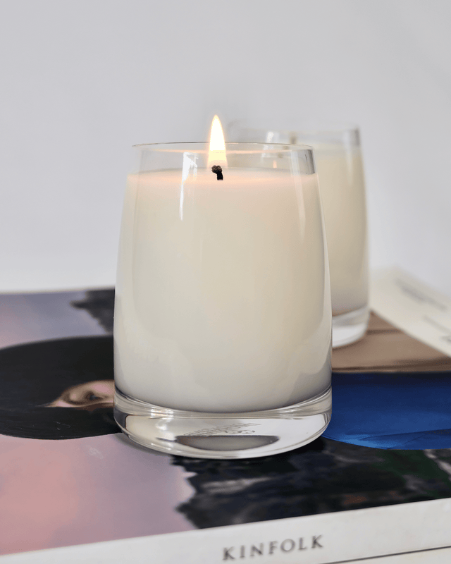 A minimalist aromatherapy candle in a clear curved glass jar, lit and displaying a soft golden flame