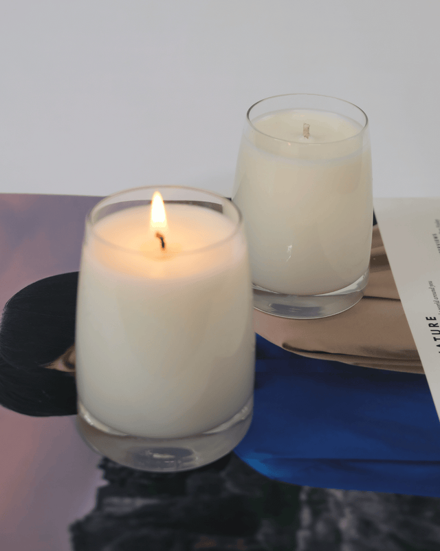Minimalist white soy wax candles in clear glass jars by Mayowa Samuel, one lit candle showing a warm flickering flame on a lifestyle magazine