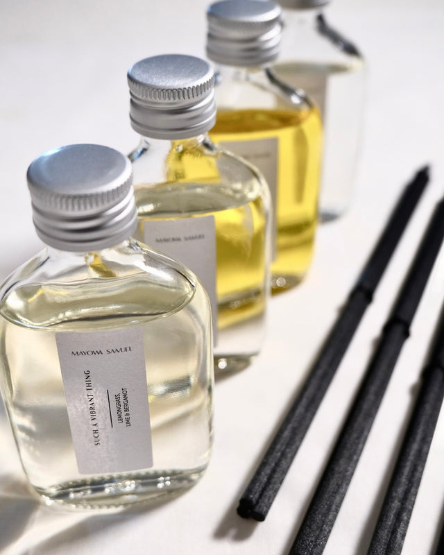 An angled, close-up product photograph of the Mayowa Samuel botanical reed diffuser range. In the sharp foreground, the "Such a Vibrant Thing" glass bottle with its silver cap and white vertical label is highly detailed, showing its clear liquid. Behind it, three other diffuser bottles recede into a soft, shallow depth of field, showcasing a gradient of clear and glowing golden oils. To the right, thick, textured black reeds run parallel, creating a clean, architectural leading line on the white surface.