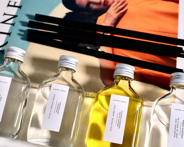An editorial, close-up lifestyle photograph of the Mayowa Samuel botanical reed diffuser collection styled elegantly across an open Kinfolk magazine. Four clear glass bottles with brushed silver metal caps sit side-by-side on a white linen surface. The clean vertical labels are highly visible, displaying "Such a Vibrant Thing" and the rich, glowing golden liquid of "Less Stress, More Zest." Sleek, textured black reeds lie horizontally in the background, completing a modern, design-focused aesthetic.