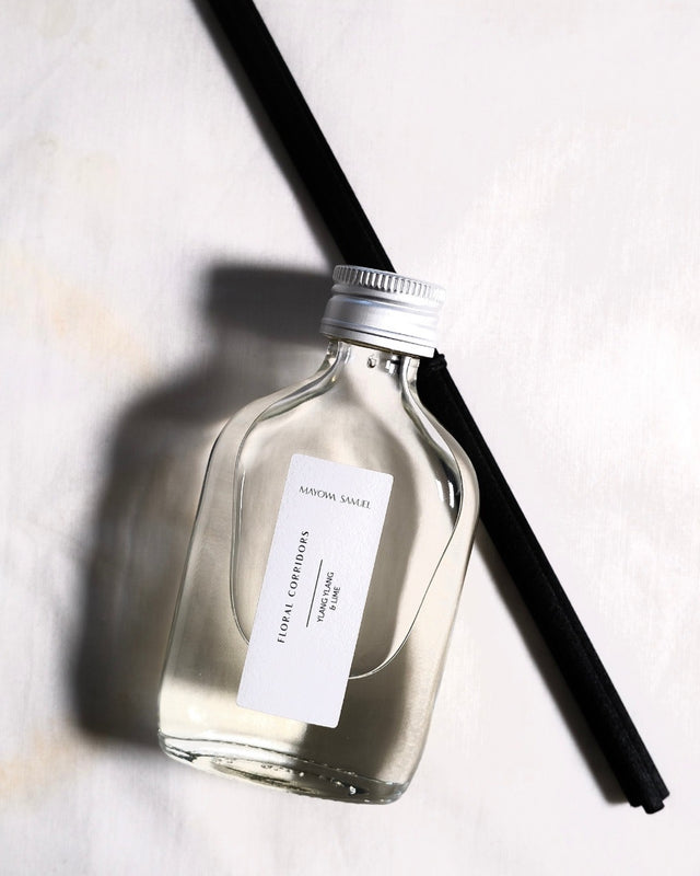 Minimalist overhead product photograph of the Mayowa Samuel "Floral Corridors" botanical reed diffuser. The clear glass bottle with a silver metal cap and a crisp white textured label rests diagonally across two sleek black reeds on a soft white fabric background, casting a soft architectural shadow.