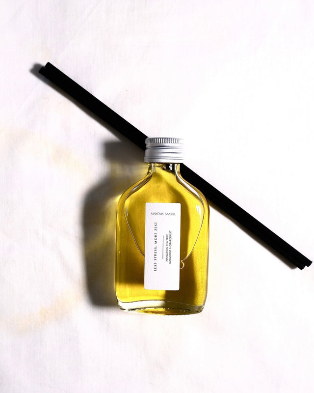 Minimalist overhead product photograph of the Mayowa Samuel "Less Stress, More Zest" botanical reed diffuser. The glass bottle contains a vibrant, glowing golden citrus oil and rests diagonally across two black reeds on a white fabric surface, casting a clean, heavy shadow to the left.