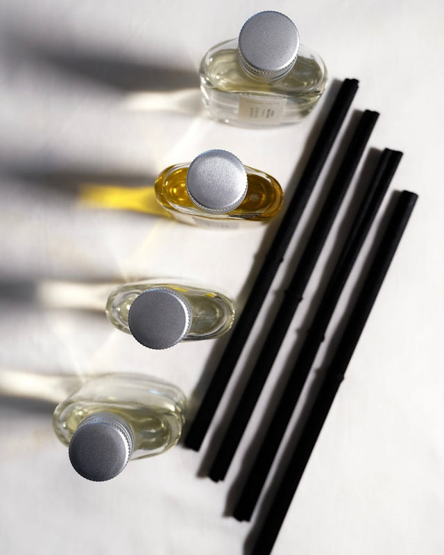 A top-down, minimalist editorial photograph of the Mayowa Samuel botanical reed diffuser collection. Four clear glass diffuser bottles with silver metal caps are arranged in a clean diagonal row on a soft textured white linen background. Next to them lie four custom, thick black reeds, creating a graphic line. Dramatic, architectural shadows stretch across the canvas from the left, highlighting the pure clarity of the liquids and the vibrant golden oil of the Less Stress, More Zest diffuser.