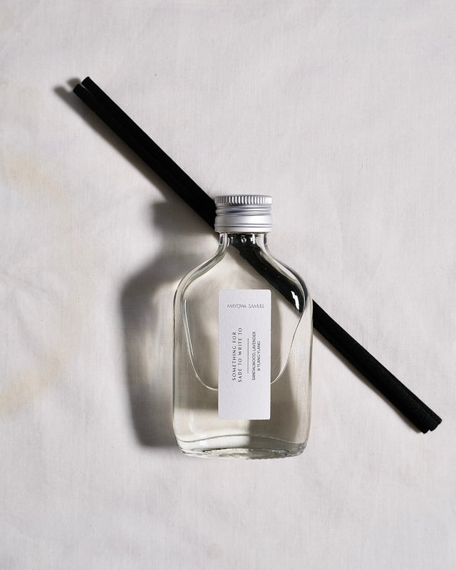 Minimalist overhead product photograph of the Mayowa Samuel "Something for Sade to write to" botanical reed diffuser. The clear glass bottle is filled with a crystal-clear liquid containing sandalwood, lavender, and ylang ylang notes. It features a brushed silver metal cap and a clean white label, resting diagonally over two textured black reeds on a soft white linen fabric background with a soft, dramatic shadow cast to the left.