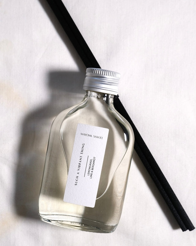 Minimalist overhead product photograph of the Mayowa Samuel "Such a Vibrant Thing" botanical reed diffuser. Featuring lemongrass, lime, and bergamot notes, the clear glass bottle with a silver metal cap rests diagonally over black reeds on a textured white linen background with elegant, soft side shadows.